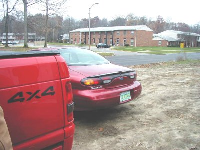 1997 Ford Taurus - A red 1997 Ford Taurus is parked next to a red pickup truck, with apartment buildings in the background.