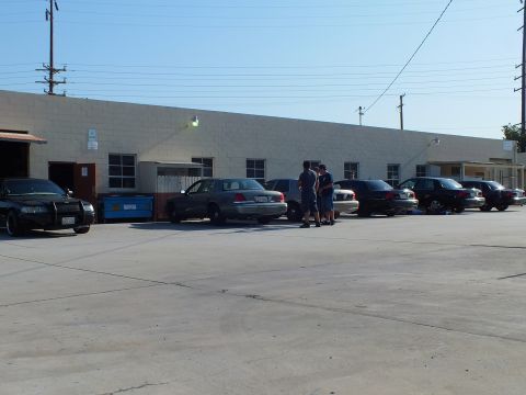2003 Ford Crown Victoria - Group of people beside a line of dark full-size sedans parked at a warehouse lot under a clear sky.
