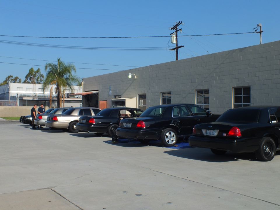 Row of black Crown Victoria sedans parked rear-out at a gray cinder-block building, one on alignment ramps