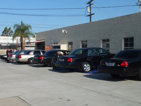 2003 Ford Crown Victoria - Mix of black and silver full-size sedans parked at a gray warehouse; one car raised on blue ramps.