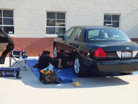 2003 Ford Crown Victoria - Two people service a black Crown Victoria LX on a tarp; PowerDirect.com decal and plate 6JBC368 on rear.