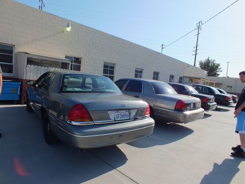 2003 Ford Crown Victoria - Silver-gray Crown Victoria parked rear-out among other sedans; plate reads 6FTU09E, a person stands at right.