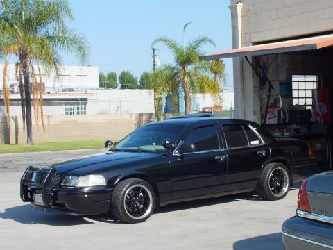 2003 Ford Crown Victoria - Lowered black Crown Victoria with brush guard and black alloy wheels outside a shop bay, palm trees behind