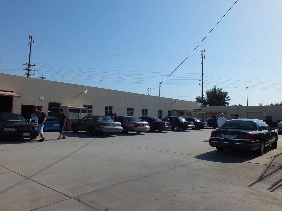 Row of sedans parked along industrial building with people gathered at car meet