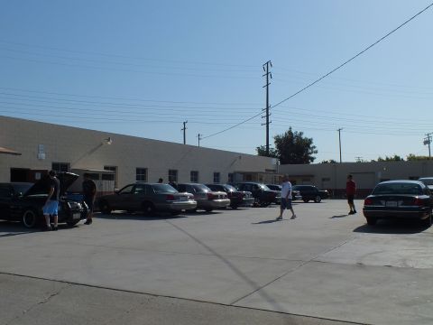 2003 Ford Crown Victoria - Wide view of a meet, sedans lined along a shop and attendees walking the lot