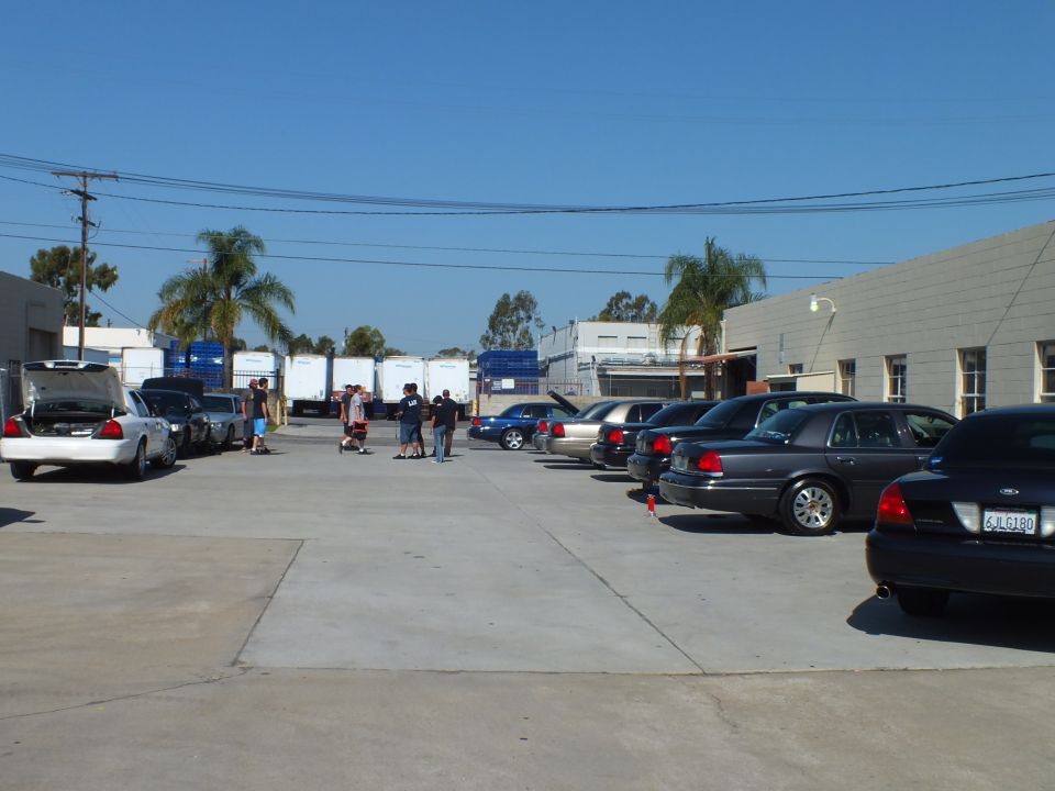 Full-size sedans parked at industrial lot with shipping containers and palms