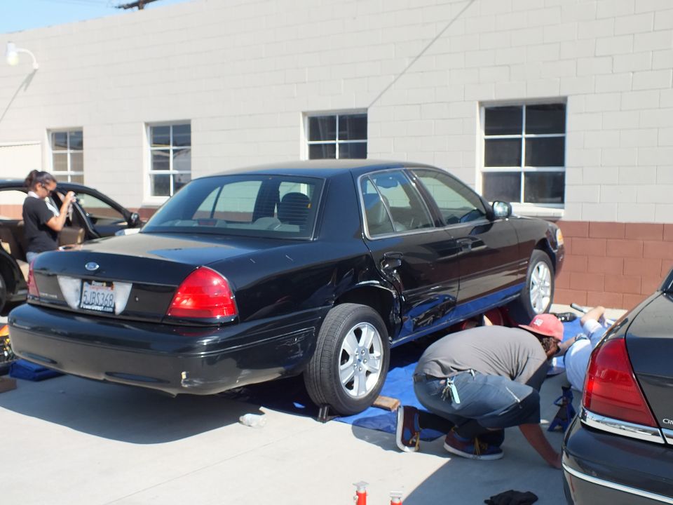 Black Ford Crown Victoria rear three-quarter with people photographing and working