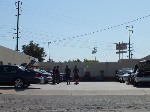 2003 Ford Crown Victoria - Several people stand between blue and silver sedans with raised hoods at a warehouse parking area.