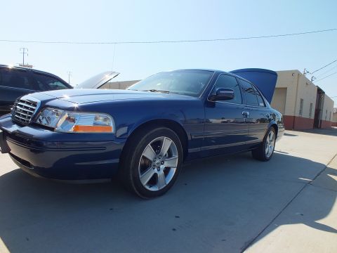 2003 Ford Crown Victoria - Dark blue full-size Ford sedan with five-spoke alloys, hood up, parked at an industrial building.