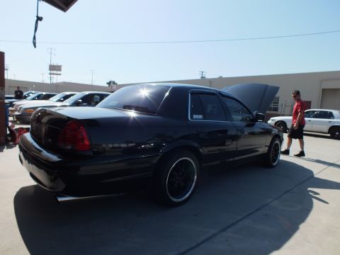 2003 Ford Crown Victoria - Black full-size sedan, trunk open, beside a person in a red shirt at a commercial lot.