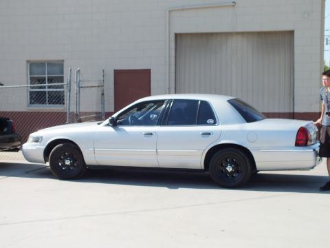 2003 Ford Crown Victoria - Silver Panther-platform sedan parked along a chain-link fence with a person standing at right.