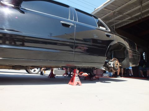 2003 Ford Crown Victoria - Side of a black sedan on jack stands, rear wheel off, as people work underneath in a shop.