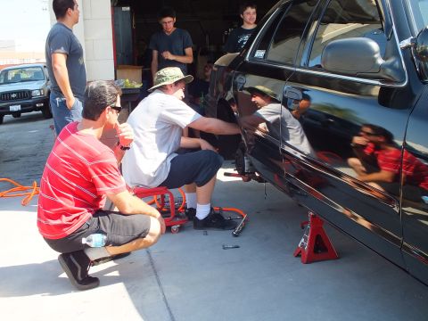 2003 Ford Crown Victoria - Several men gather around a black sedan on jack stands during a rear suspension repair.