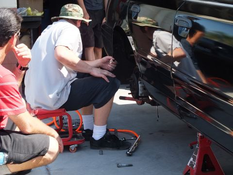 2003 Ford Crown Victoria - Attendees sit on a creeper next to a black car on stands, hand tools laid on the concrete.