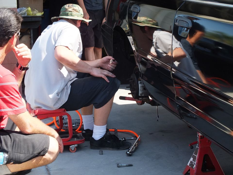 Men crouched beside black sedan on jack stands working with creeper and tools
