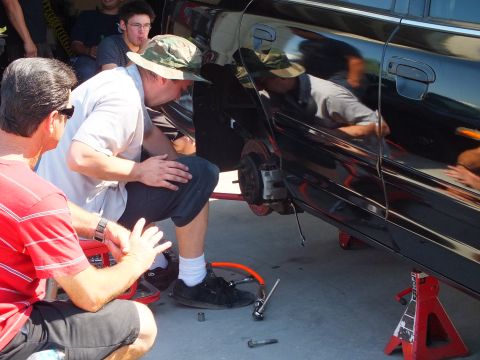 2003 Ford Crown Victoria - Group inspects the bare rear hub and rotor of a black sedan supported on red jack stands.