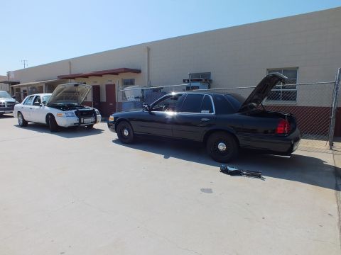 2003 Ford Crown Victoria - White Ford sedan with hood up next to a black Crown Victoria with open trunk at a shop lot.
