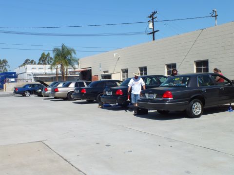 2003 Ford Crown Victoria - Attendees stand by a line of dark Ford sedans parked outside an industrial warehouse.