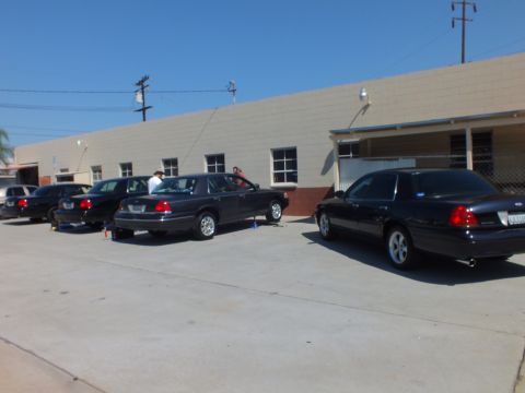 2003 Ford Crown Victoria - Several dark full-size Ford sedans lined up beside a warehouse under a clear blue sky.