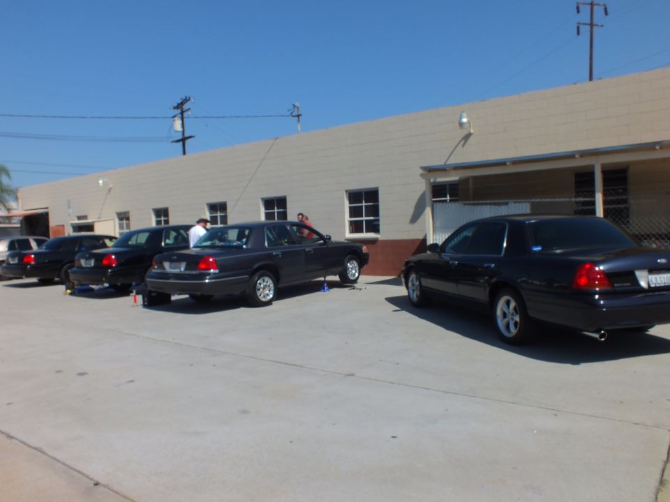 Row of dark Crown Victoria sedans parked along beige industrial building with palm trees