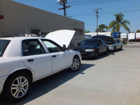 2003 Ford Crown Victoria - White full-size sedan, hood raised, parked ahead of black and silver Fords at a shop lot.