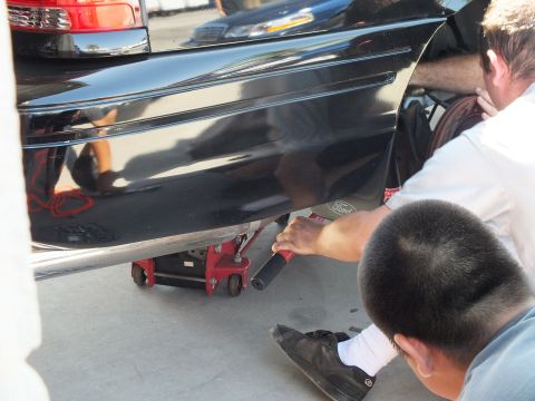 2003 Ford Crown Victoria - Worker slides a red low-profile floor jack beneath the black sedan's rocker, Ford emblem mud flap visible.