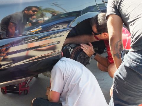 2003 Ford Crown Victoria - Sunlit shop lot: men examine the rear hub and rotor of a lowered black Ford sedan, glossy body reflecting bystanders.