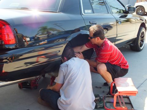 2003 Ford Crown Victoria - Mechanics work the rear corner of a black Crown Victoria; red rolling tool cart and floor jack sit on the pavement.