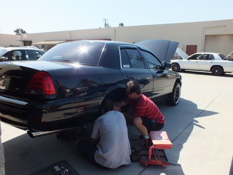2003 Ford Crown Victoria - Rear-three-quarter view of a black Crown Victoria, hood raised, as two men service the back wheel; white sedans behind.