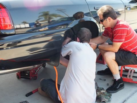 2003 Ford Crown Victoria - Kneeling helper and seated man in red work the bare rear hub of a black Ford sedan; orange air hose and sockets on ground.
