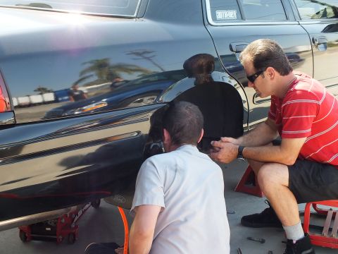 2003 Ford Crown Victoria - Mechanics crouch at the open rear fender of a black Ford sedan, hub exposed, hardware laid out on the concrete.