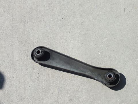 2003 Ford Crown Victoria - removed black stamped-steel rear lower control arm with pressed-in bushings rests on a sunlit concrete floor.