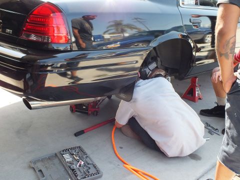 2003 Ford Crown Victoria - Mechanic lies at the rear wheel of a black Crown Victoria on stands; an open socket tray and orange hose sit on the lot.