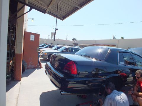 2003 Ford Crown Victoria - Rear view of a black Crown Victoria beside a row of parked sedans under a shop overhang; tires stacked at left.