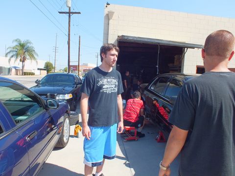 2003 Ford Crown Victoria - Bystander in a graphic tee faces the camera while crew works a black sedan and a blue sedan sits at the shop driveway.