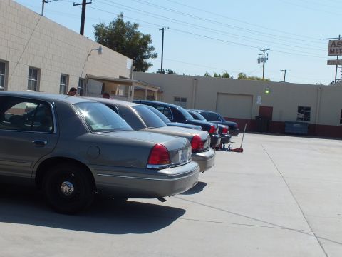 2003 Ford Crown Victoria - Several Panther-platform Ford sedans line up in a paved lot beside an industrial building under clear sky.