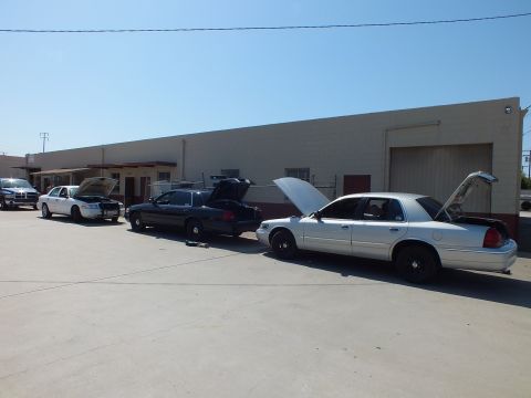 2003 Ford Crown Victoria - White, dark and silver Panther-platform sedans sit with hoods up outside a beige industrial shop in a paved lot.