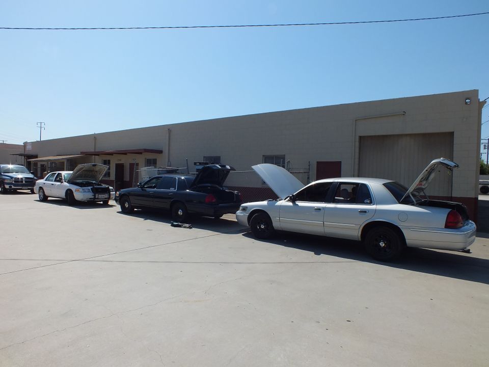 Three Ford Crown Victoria sedans with hoods and trunks open parked outside shop building