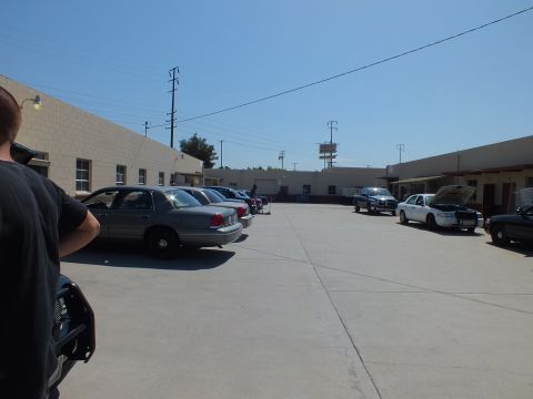2003 Ford Crown Victoria - Wide view of a shop yard: sedans line the left building wall while more cars park across the open concrete lot.
