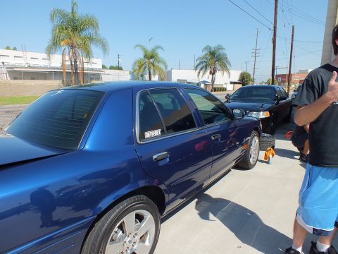2003 Ford Crown Victoria - Side view of a blue Crown Victoria raised on a jack near the shop, chrome alloy wheels and tinted glass catching light.