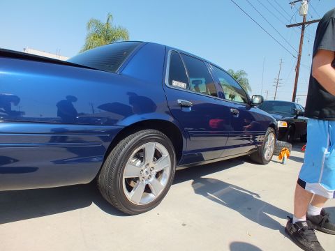 2003 Ford Crown Victoria - Blue Crown Victoria rear corner raised on a yellow-handled jack, chrome alloys gleaming; a man stands at right.