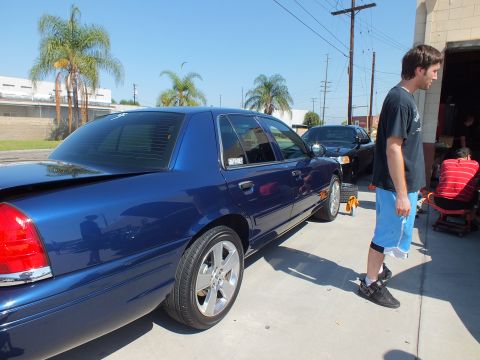 2003 Ford Crown Victoria - Side profile of a blue Crown Victoria raised at the shop driveway while a bystander looks on; palms line the background.