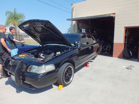 2003 Ford Crown Victoria - Black Crown Victoria with police-style push bar sits hood-open on yellow ramps; two men stand at left of the shop.