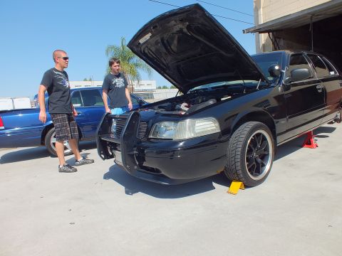 2003 Ford Crown Victoria - Black Crown Victoria with brush guard sits on ramps, hood up; two men in tees stand at the front fender near the shop.