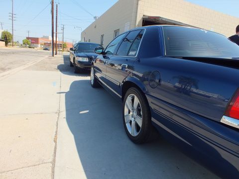 2003 Ford Crown Victoria - Rear three-quarter view of a navy full-size Ford sedan parked along a concrete drive beside a tan warehouse.
