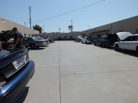 2003 Ford Crown Victoria - Row of full-size sedans parked between warehouses, foreground car hood raised for service.