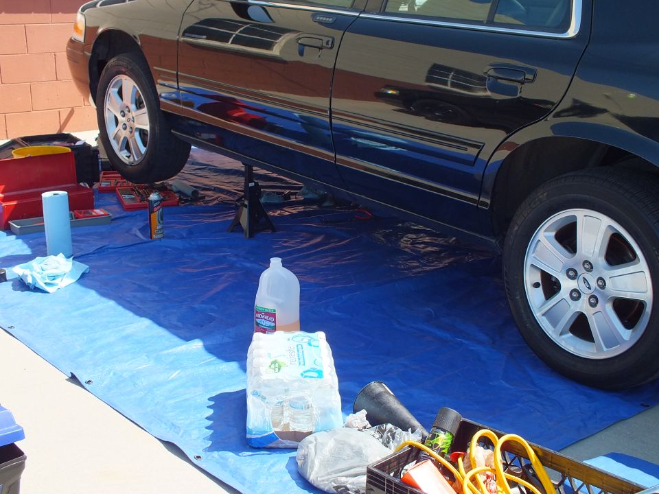 Black Ford sedan raised on jack stands over a blue tarp with tools and coolant jug