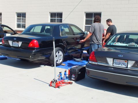 2003 Ford Crown Victoria - Workers stand between two black full-size Fords with a red floor jack and toolbox on the pavement.