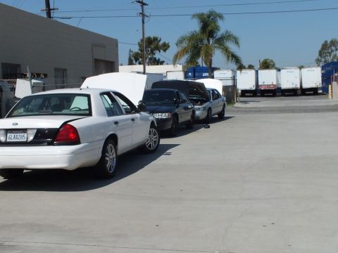 2003 Ford Crown Victoria - White full-size Ford parked ahead of darker sedans near rows of white freight trailers.