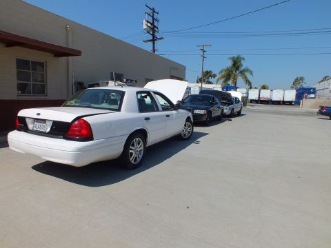 2003 Ford Crown Victoria - White Panther sedan, hood raised, leading a line of cars along a warehouse and freight trailers.